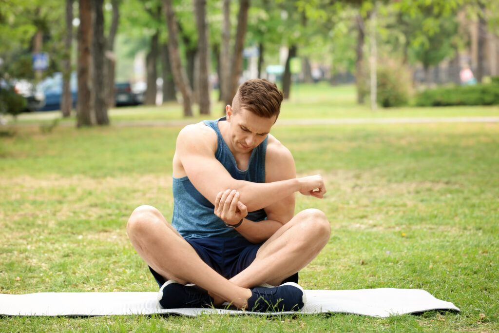 Male athlete sitting on ground outdoors holding his painful elbow after training, sports injury care at Mast Health in Tampa