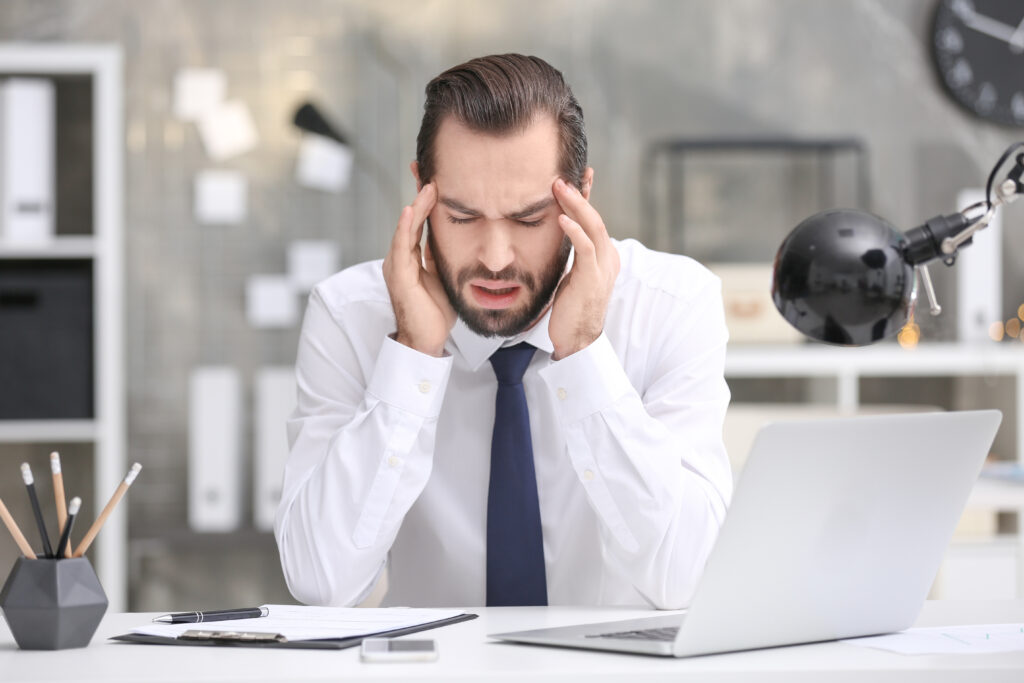 Man in dress shirt pressing his temples in pain at his office desk, tension headache relief Mast Health offers Tampa professionals