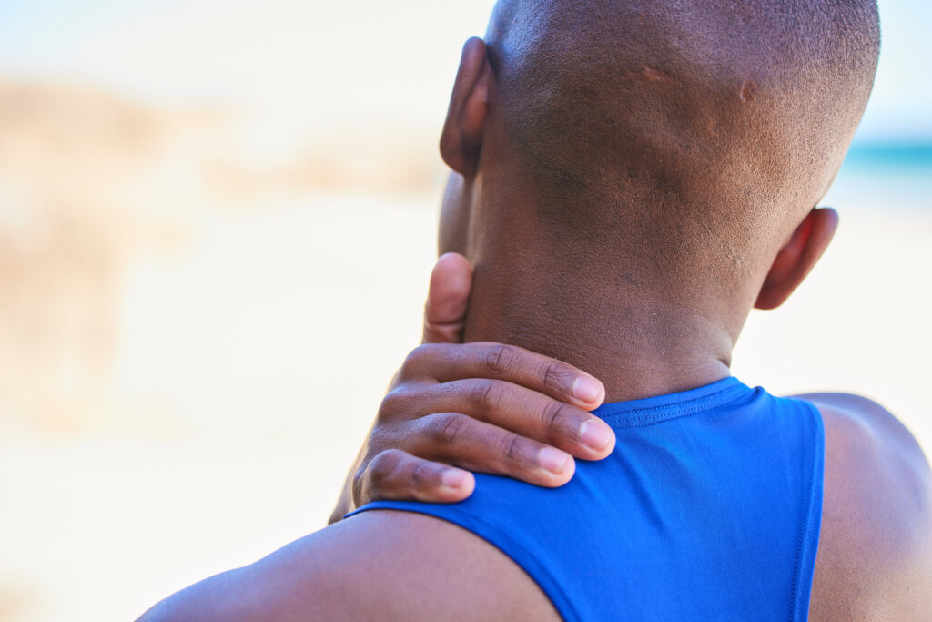 Man standing outdoors at the beach gripping the back of his neck in pain after exercise, a common neck strain treated at Mast Health in Tampa, FL