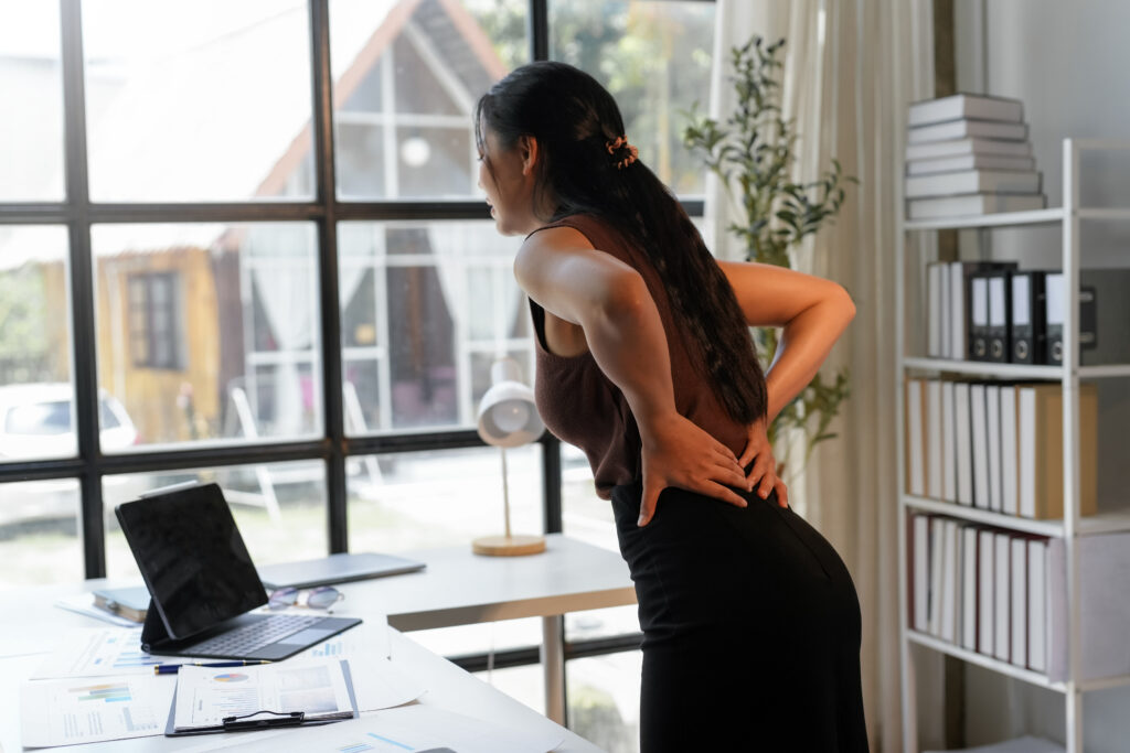 Businesswoman standing at her desk arching backward and holding her lower back in pain, posture-related back pain treated at Mast Health in Tampa, FL
