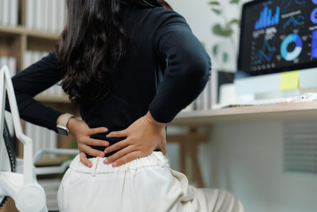 Businesswoman holding her lower back in pain at her office desk, sciatica relief provided at Mast Health chiropractic clinic in Tampa, FL