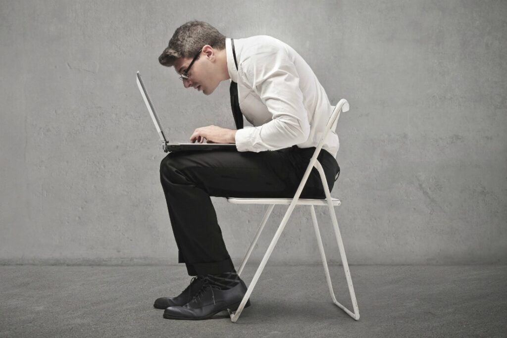 Man sitting on a stool with his back rounded and head dropped forward over a laptop, poor posture corrected at Mast Health in Tampa, FL