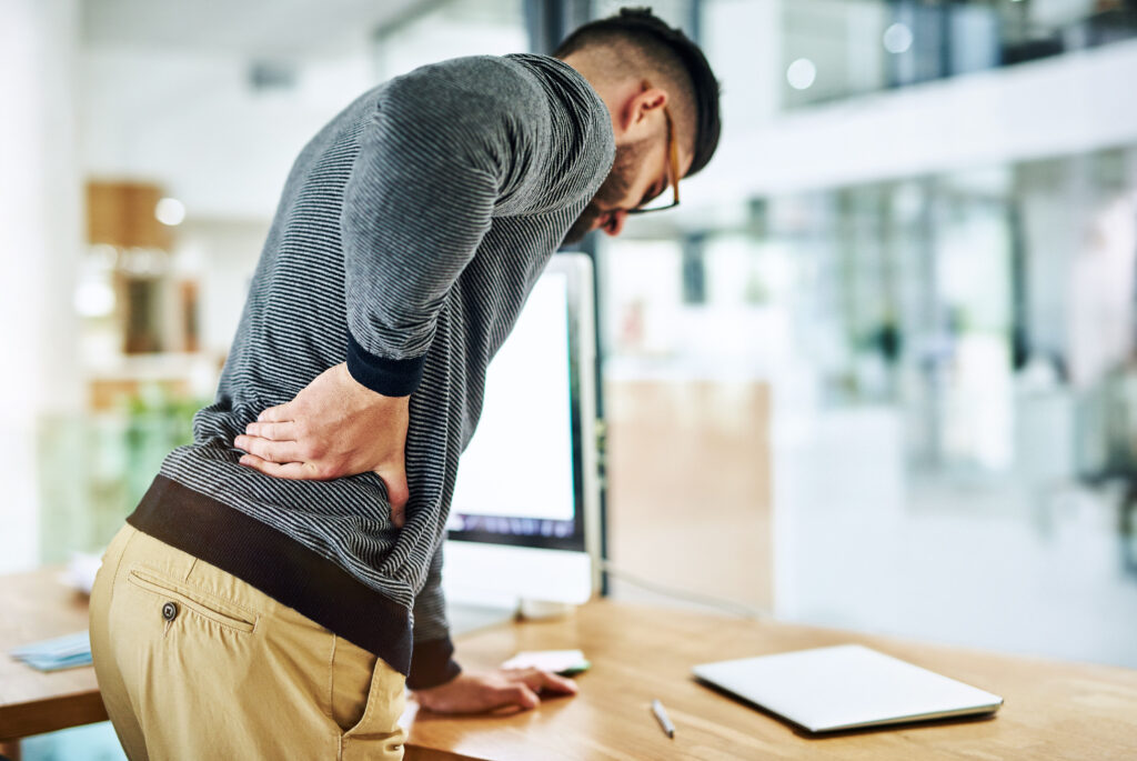 Man bent over holding his lower back in pain at his office desk, back pain Mast Health treats for Tampa professionals