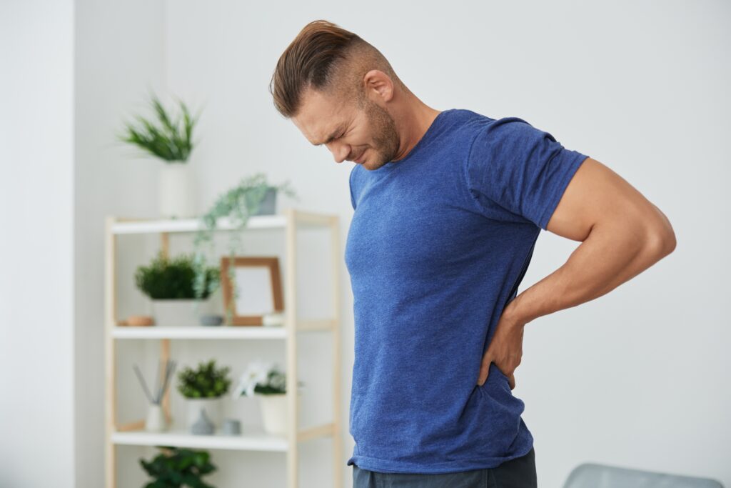 Man in blue shirt bent forward holding his neck and shoulder in pain at home, back pain and muscle strain Mast Health treats in Tampa