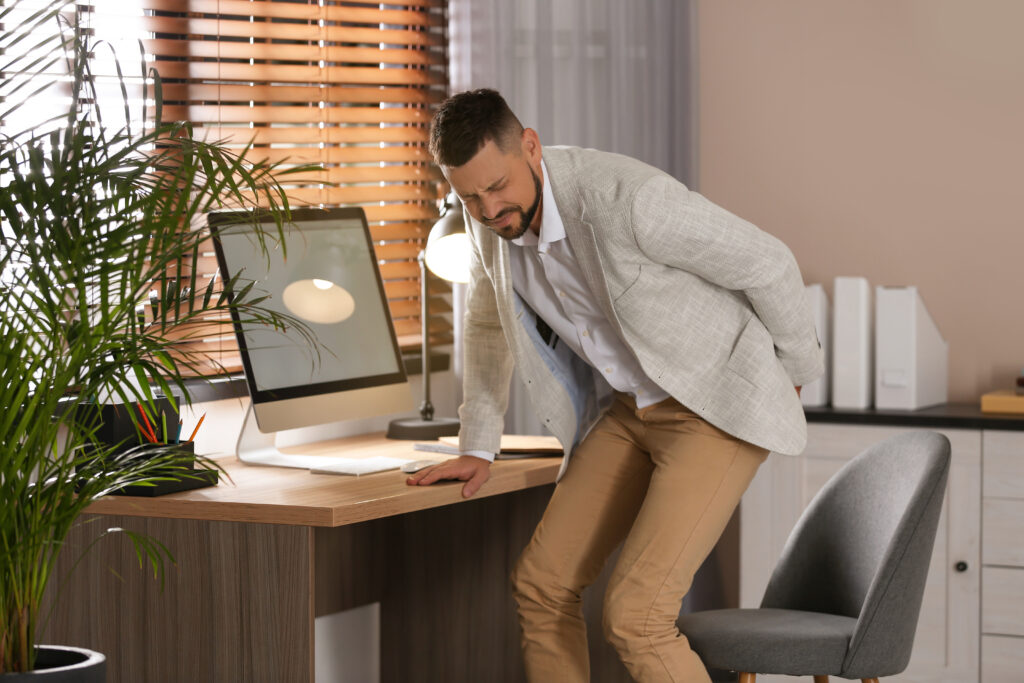 Man leaning heavily over his office desk with one hand braced on it, showing signs of discomfort from poor posture at work, care available at Mast Health in Tampa, FL