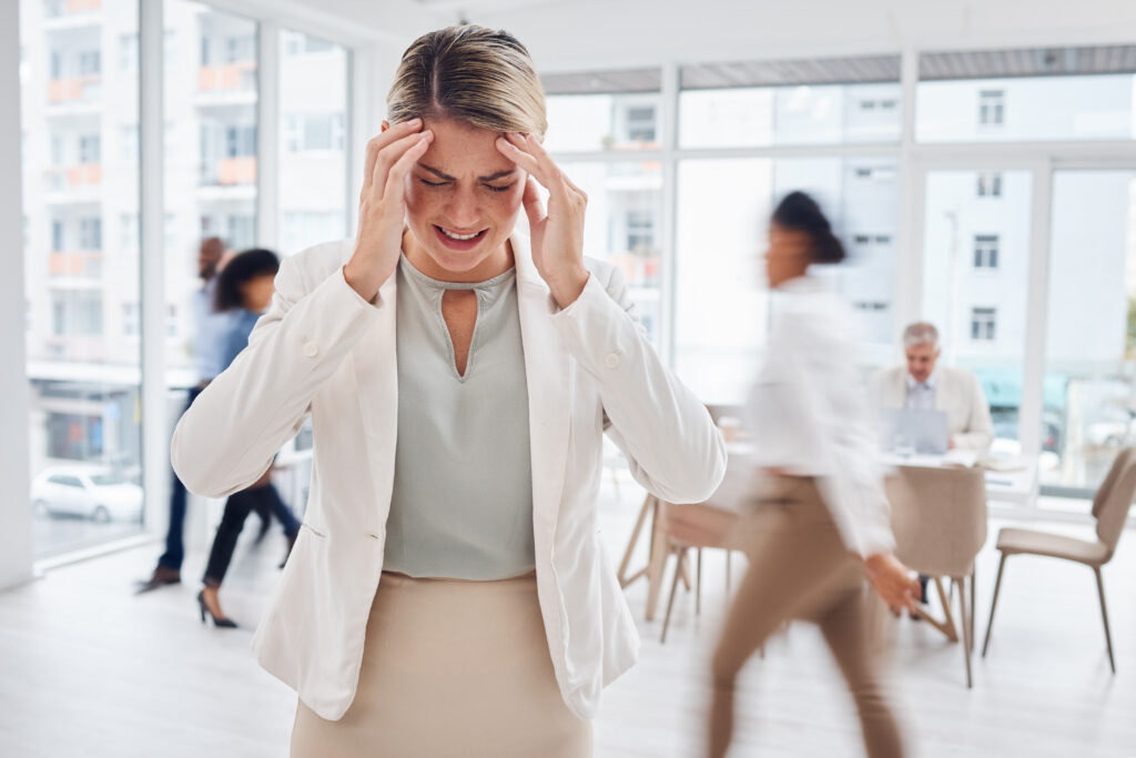 Businesswoman gripping her head in pain amid a busy office, a sign of vertigo and dizziness treated at Mast Health in Tampa, FL