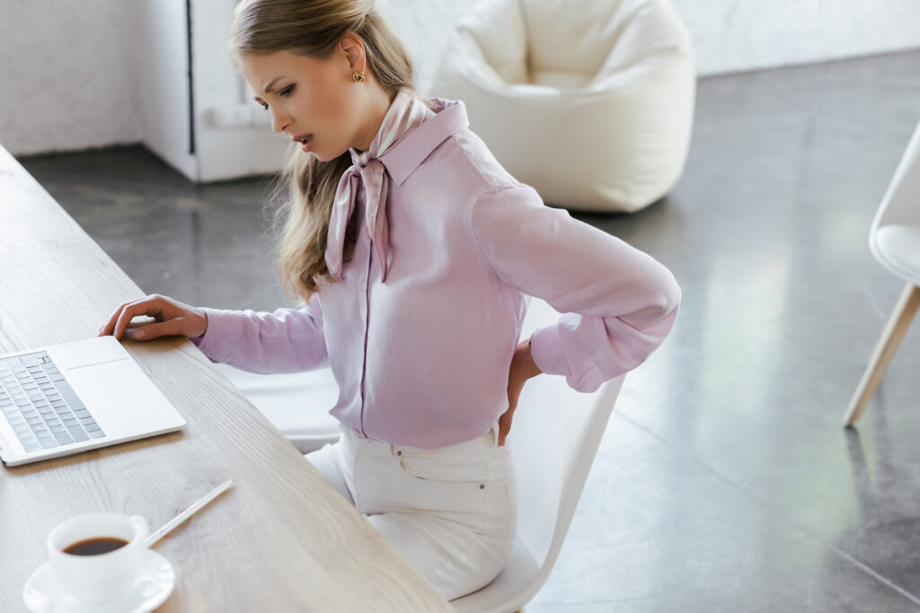 Businesswoman leaning over her laptop at a counter-height desk while touching her back in pain, posture-related back strain treated at Mast Health in Tampa, FL