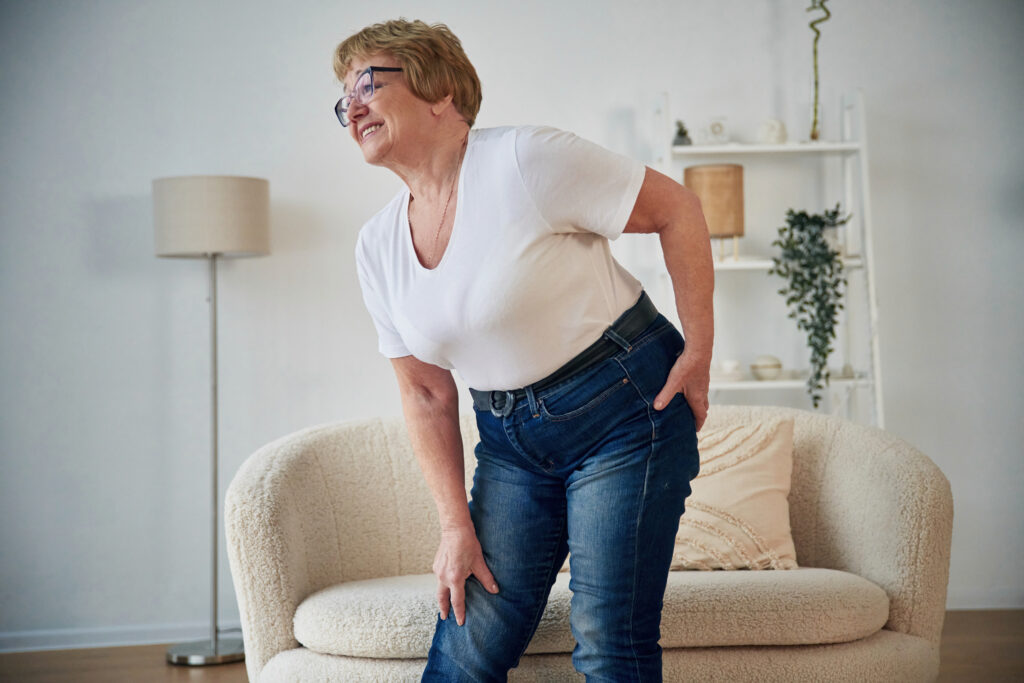 Senior woman standing in her living room holding her lower back and hip in discomfort, a common sign of hip pain treated at Mast Health in Tampa, FL