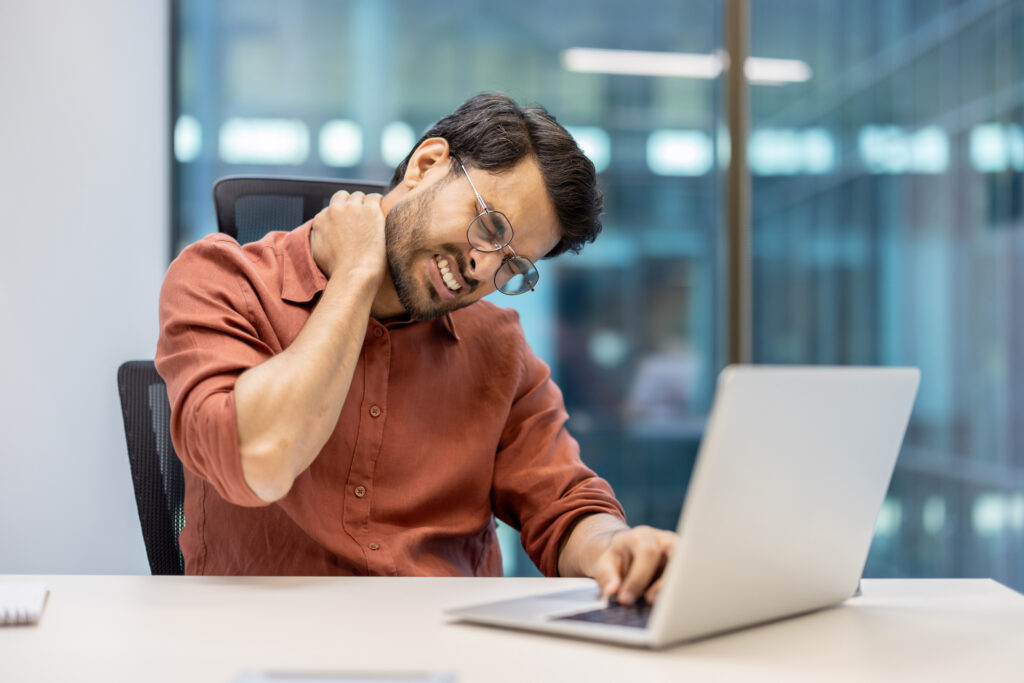 Office worker wincing in pain and holding the back of his neck while working on his laptop, a common office-related neck pain symptom treated at Mast Health in Tampa, FL