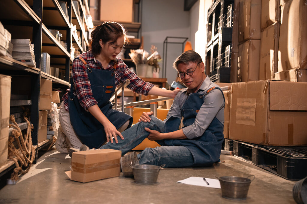 A coworker helps examine a warehouse worker's knee after a workplace injury, an overuse injury treated at Mast Health in Tampa, FL