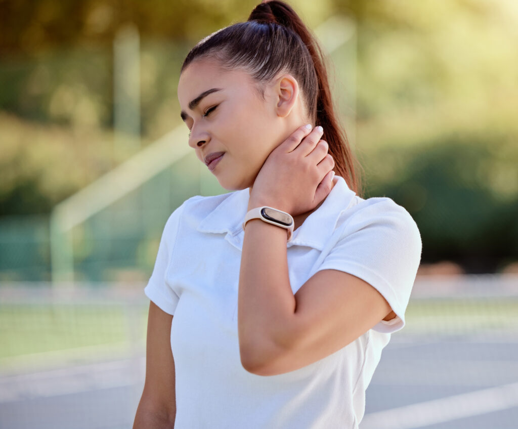 Young female tennis athlete outdoors on the court gripping her neck in discomfort during a match, a common sports-related neck pain treated at Mast Health in Tampa, FL