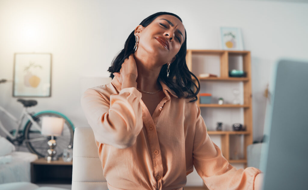 Woman tilting her head back with eyes closed while gripping the side of her neck in pain at her home office desk, a symptom of neck pain treated at Mast Health in Tampa, FL